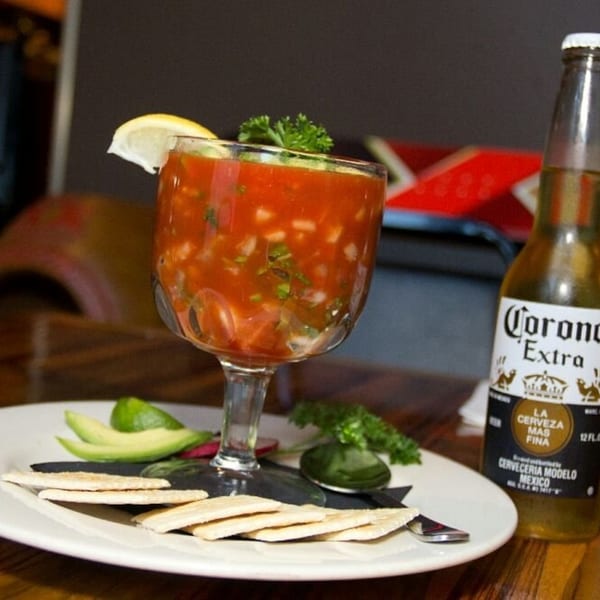 A glass of red ceviche with a lemon slice, avocado, crackers, and a Corona beer.