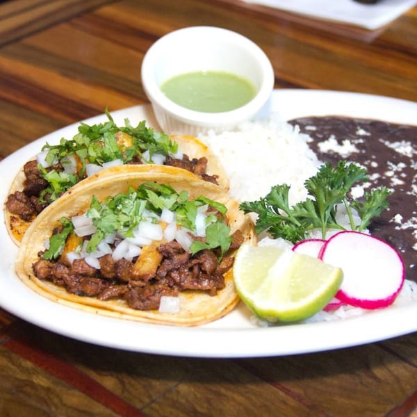 Two tacos with meat, cilantro, and onions, served with rice, black beans, lime, and radishes.